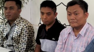 Tiga orang pria duduk di meja sambil melihat ke arah kamera