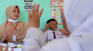 Seorang guru perempuan sedang menjelaskan materi pelajaran kepada siswa di kelas.