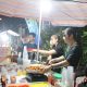 Seorang penjual sedang menggoreng makanan di sebuah stand makanan di Bazar Kuliner Kampoeng Cungking. Terlihat beberapa orang pengunjung yang antusias mendekat ke stand tersebut.