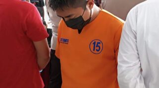 Seorang pria mengenakan baju tahanan berwarna orange sedang berdiri dengan tangan diborgol.