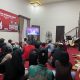 Sejumlah orang sedang menonton televisi di sebuah ruangan. Suasana terlihat ramai dan hangat.