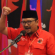 Seorang pria berbaju merah sedang berorasi dengan gestur semangat, sambil memegang mikrofon.