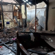 Foto menunjukkan sisa-sisa bangunan rumah yang hangus terbakar. Terlihat puing-puing kayu dan atap yang runtuh.