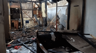 Foto menunjukkan sisa-sisa bangunan rumah yang hangus terbakar. Terlihat puing-puing kayu dan atap yang runtuh.