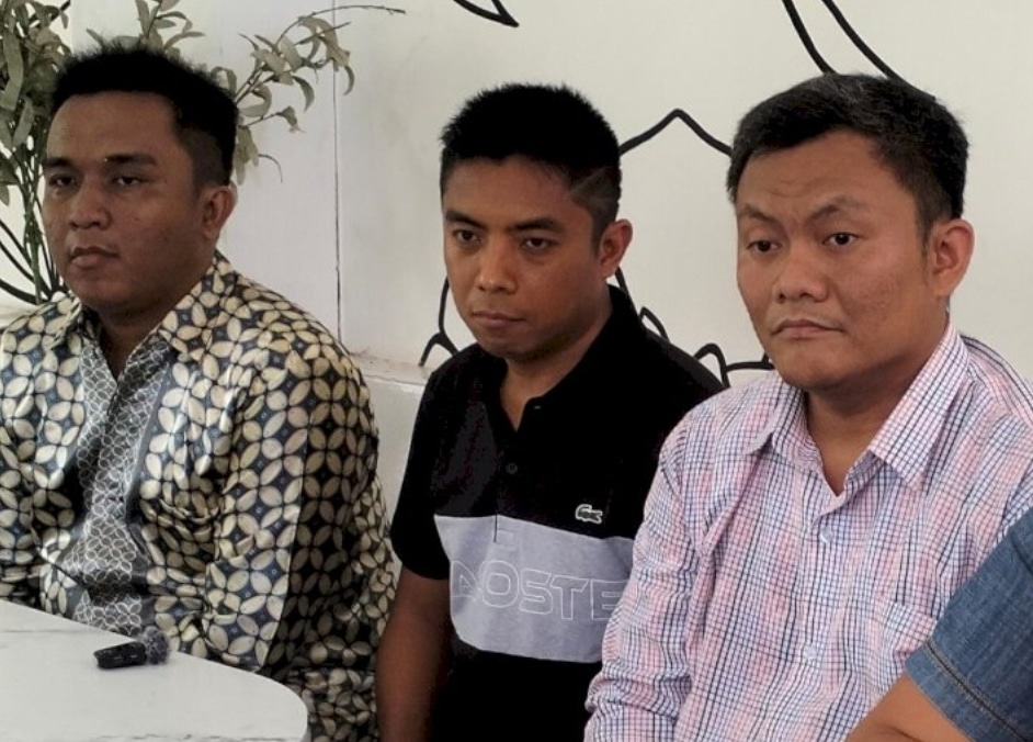 Tiga orang pria duduk di meja sambil melihat ke arah kamera