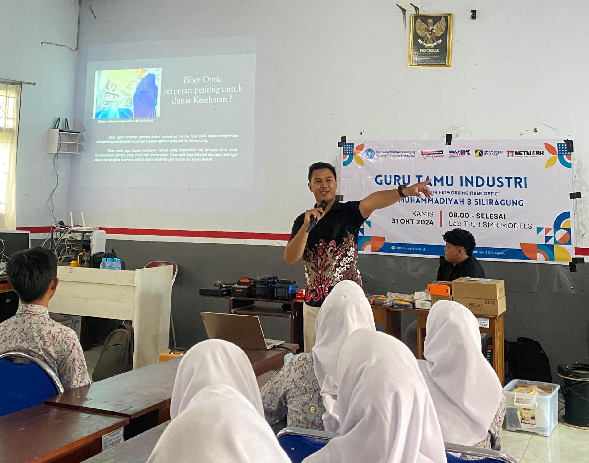 Foto menunjukkan seorang instruktur sedang menjelaskan materi fiber optik di depan para siswa di SMK Models.