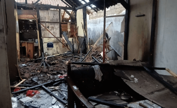 Foto menunjukkan sisa-sisa bangunan rumah yang hangus terbakar. Terlihat puing-puing kayu dan atap yang runtuh.