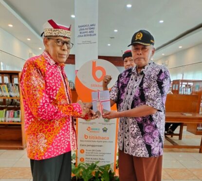 IGB Sudharma tersenyum memegang buku, mengenakan udeng merah putih, didampingi petugas perpustakaan.